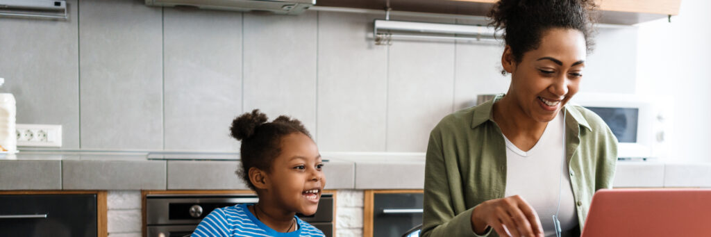 Woman working with laptop while sitting at table with her daughter in home kitchen